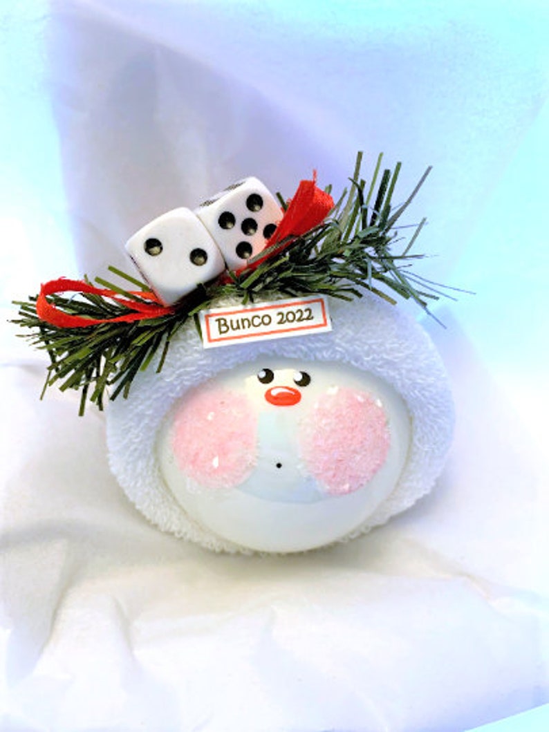 May include: A white snowman ornament with pink cheeks, a red nose, and black eyes. The ornament is topped with two dice, green sprigs, and a red ribbon. A small tag reads "Bunco 2022".