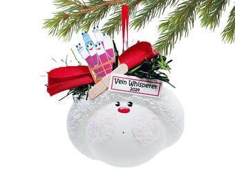 May include: A white snowman-shaped Christmas ornament with a red nose and rosy cheeks. It features a bandage, a red ribbon, and a sign that reads "Vein Whisperer 2024." The ornament also includes a depiction of cartoon veins.