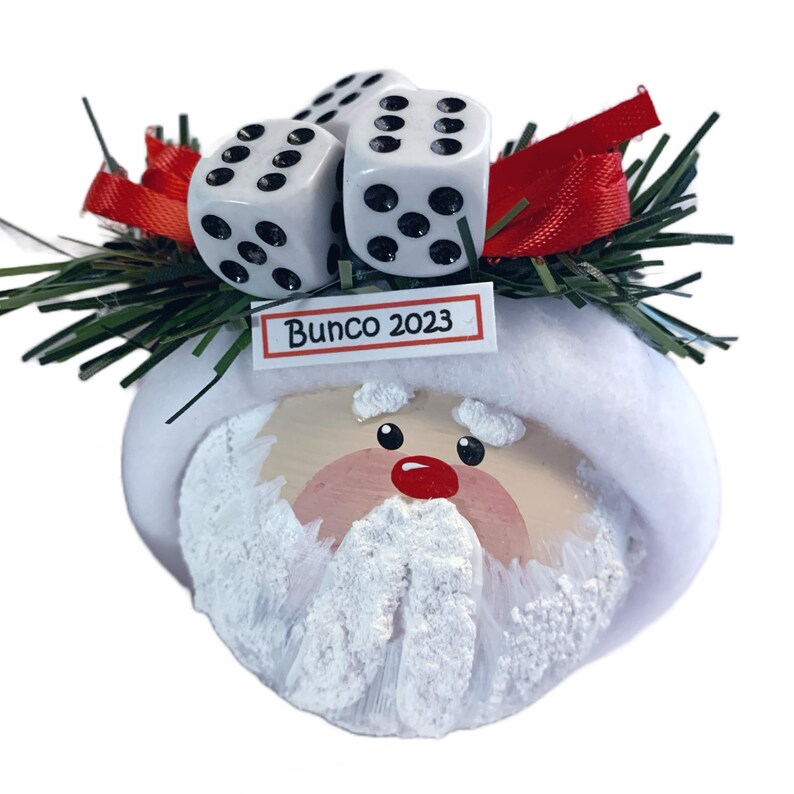 May include: A round Santa Claus ornament featuring a white fuzzy hat brim, a painted face with a red nose, and a textured white beard. The top is decorated with green pine needles, red ribbon, three white dice, and a tag reading Bunco 2023.