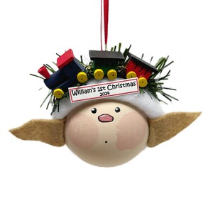 May include: A wooden elf ornament with a white fur hat and brown felt ears. The elf is wearing a toy train on its head with a red, blue, and yellow train car. The ornament has a white banner that reads "William's 1st Christmas 2024".