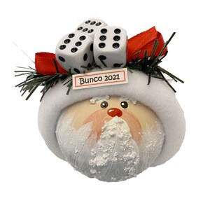 May include: A round Christmas ornament featuring a painted Santa Claus face with a white fuzzy hat brim. Three white dice with black pips are attached to the top, accented by a red ribbon and green pine needles. A small sign reads Bunco 2021.
