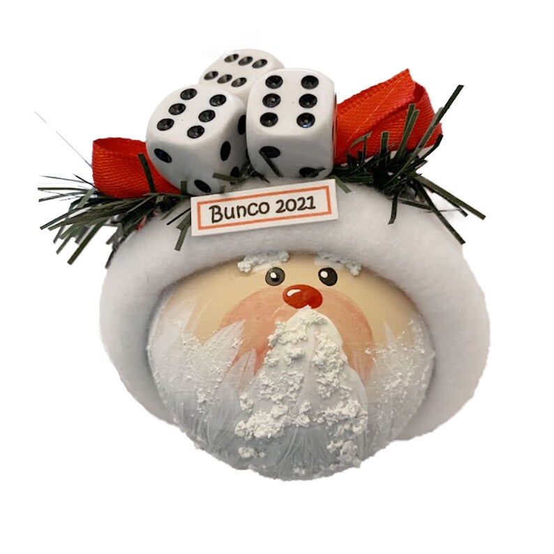 May include: A round Christmas ornament featuring a painted Santa Claus face with a white fuzzy hat brim. Three white dice with black pips are attached to the top, accented by a red ribbon and green pine needles. A small sign reads Bunco 2021.
