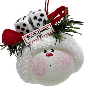 May include: A white ornament with a snowman face, pink cheeks, and a red mouth. It has a white hat, two dice, a red bow, and the text "Bunco Queen 2025". The ornament is decorated with green sprigs.