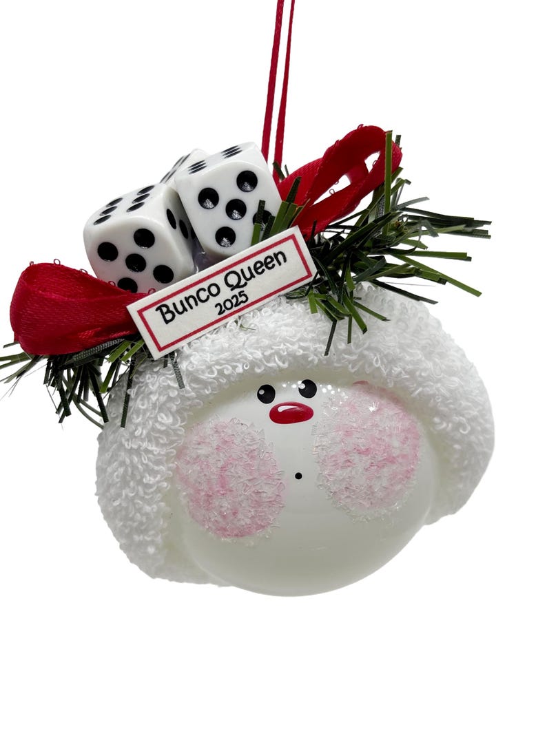 May include: A white ornament with a snowman face, pink cheeks, and a red mouth. It has a white hat, two dice, a red bow, and the text "Bunco Queen 2025". The ornament is decorated with green sprigs.