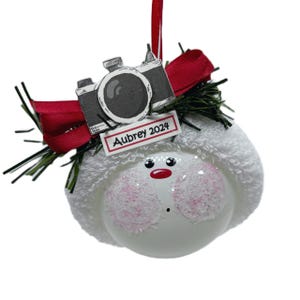 May include: A white Christmas ornament with a snowman face, pink glitter cheeks, and a red nose. It features a camera and a tag that reads "Aubrey 2024". The ornament is decorated with a red bow and green sprigs.