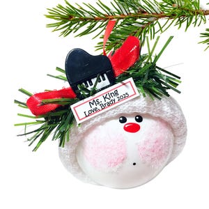 Puede incluir: Adorno navideño con forma de cabeza de muñeco de nieve, con gorro blanco de punto. Tiene un piano negro y una etiqueta que dice "Ms. King Love, Brady 2025". El muñeco de nieve tiene mejillas rosas, nariz roja y ojos negros.