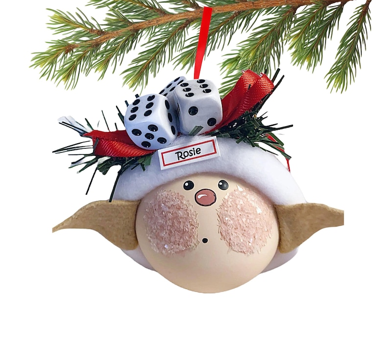 Puede incluir: Adorno navide&ntilde;o caprichoso con una cara redonda, mejillas rosadas, un sombrero blanco y orejas beige. Dos dados blancos con puntos negros est&aacute;n encima del sombrero, con una cinta roja y el nombre "Rosie".
