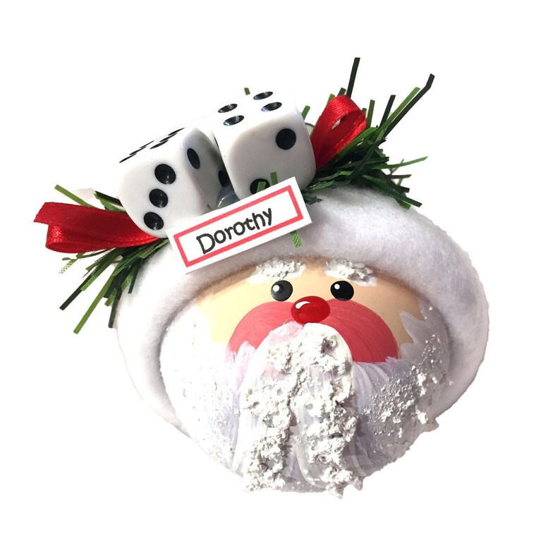 May include: A round Christmas ornament shaped like a Santa Claus face with a white plush hat brim. It features two white dice, green pine needles, and a red ribbon on top. A small white rectangular tag with a red border displays the name Dorothy.
