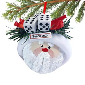 May include: A round Santa Claus ornament featuring a white hat, a textured white beard, and a red nose. The top is decorated with green pine needles, red ribbon, and three white dice with black pips. A small white sign reads Bunco 2023.