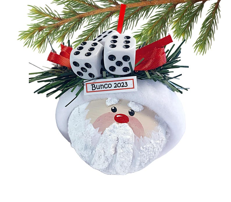 May include: A round Santa Claus ornament featuring a white hat, a textured white beard, and a red nose. The top is decorated with green pine needles, red ribbon, and three white dice with black pips. A small white sign reads Bunco 2023.
