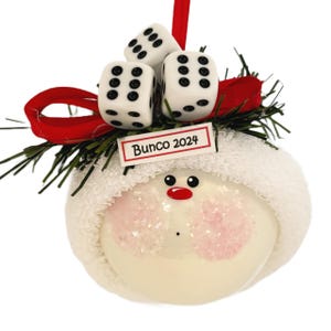 May include: A festive Christmas ornament with a snowman design. The ornament features a white face with pink cheeks, a red nose, and a white hat. Three dice are on top, and the text "Bunco 2024" is displayed on a small sign.