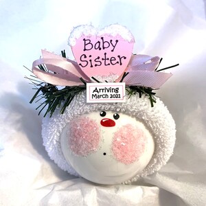 Puede incluir: Adorno navideño blanco con un corazón rosa que dice "Baby Sister" y una etiqueta que dice "Arriving March 2021".