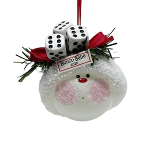 May include: A white ornament with a round, fluffy head, pink cheeks, and a red nose. Three white dice with black dots sit atop a red bow and green sprigs. The ornament has a tag that reads "Bunco Babe 2024".
