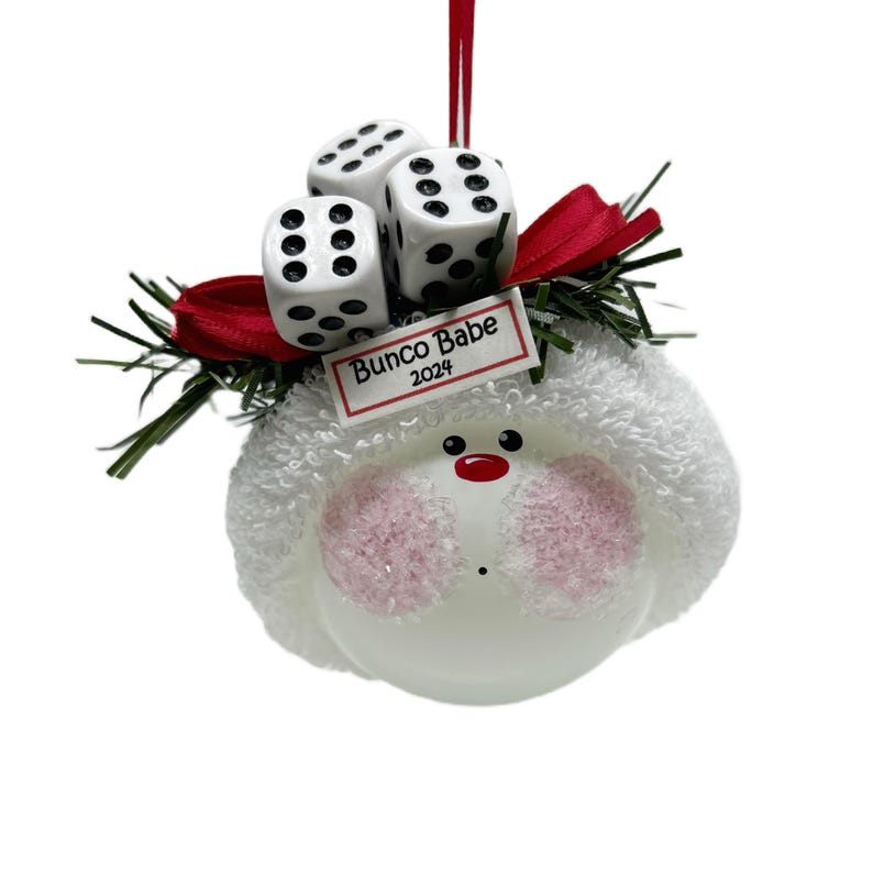 May include: A white ornament with a round, fluffy head, pink cheeks, and a red nose. Three white dice with black dots sit atop a red bow and green sprigs. The ornament has a tag that reads "Bunco Babe 2024".