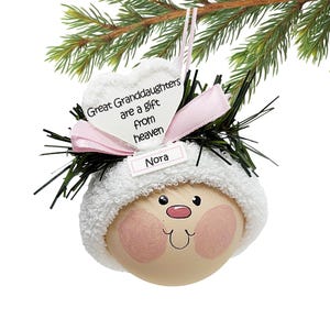 May include: A Christmas ornament featuring a painted face with rosy cheeks, a white hat, and a pink bow. The ornament has a heart-shaped tag that reads "Great Granddaughters are a gift from heaven" and a name tag that says "Nora".