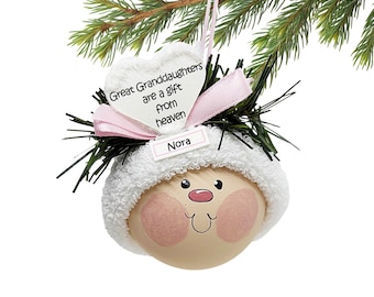May include: A Christmas ornament featuring a painted face with rosy cheeks, a white hat, and a pink bow. The ornament has a heart-shaped tag that reads "Great Granddaughters are a gift from heaven" and a name tag that says "Nora".