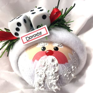 May include: A round Christmas ornament shaped like a Santa Claus face with a white felt hat brim, a red nose, and a textured white beard. Two white dice with black pips, green pine needles, and a red ribbon sit on top. A white tag reads Dorothy.