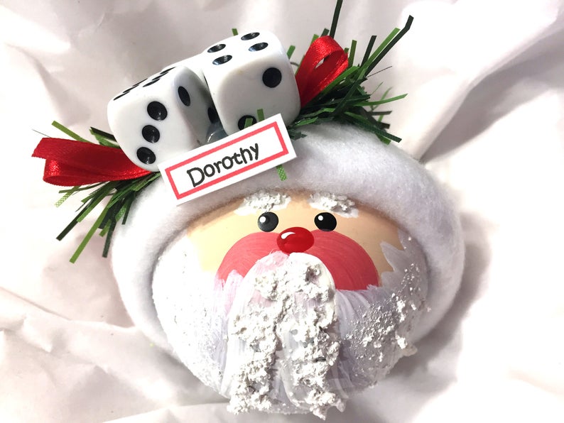 May include: A round Christmas ornament shaped like a Santa Claus face with a white felt hat brim, a red nose, and a textured white beard. Two white dice with black pips, green pine needles, and a red ribbon sit on top. A white tag reads Dorothy.