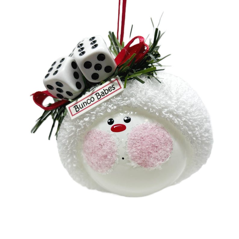 May include: A white ornament with a snowman face, pink cheeks, and a red nose. Two dice and a tag that reads "Bunco Babes" are attached to the top with red ribbon and green sprigs. A festive holiday decoration.