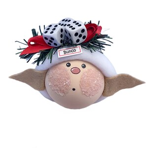 Puede incluir: Un adorno caprichoso con una cara redonda y clara, mejillas rosadas y una nariz peque&ntilde;a. Tiene un sombrero blanco, alas color canela y est&aacute; decorado con dos dados y un lazo rojo. La palabra "Bunco" est&aacute; impresa en una peque&ntilde;a etiqueta.