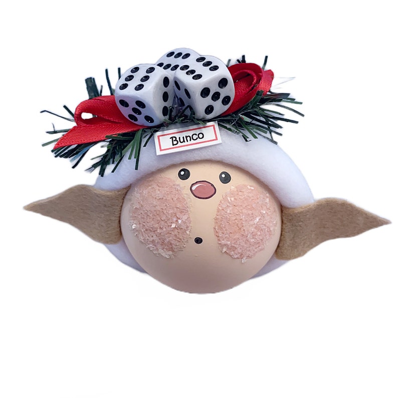 Puede incluir: Un adorno caprichoso con una cara redonda y clara, mejillas rosadas y una nariz peque&ntilde;a. Tiene un sombrero blanco, alas color canela y est&aacute; decorado con dos dados y un lazo rojo. La palabra "Bunco" est&aacute; impresa en una peque&ntilde;a etiqueta.