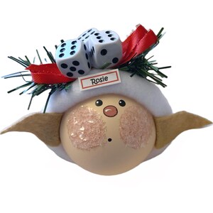 Puede incluir: Un adorno navide&ntilde;o caprichoso con una cara redonda y clara, mejillas rosadas y una boca peque&ntilde;a y sonriente. El adorno tiene un sombrero blanco, orejas color canela y est&aacute; adornado con dos dados y una cinta roja. El nombre "Rosie" est&aacute; impreso en una peque&ntilde;a etiqueta.