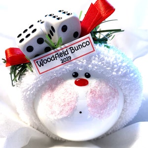 May include: A white ornament with a snowman face, pink cheeks, and a red nose. Two dice sit atop the ornament, along with a tag that reads "Woodfield Bunco 2019". The ornament is decorated with red ribbon and green sprigs.