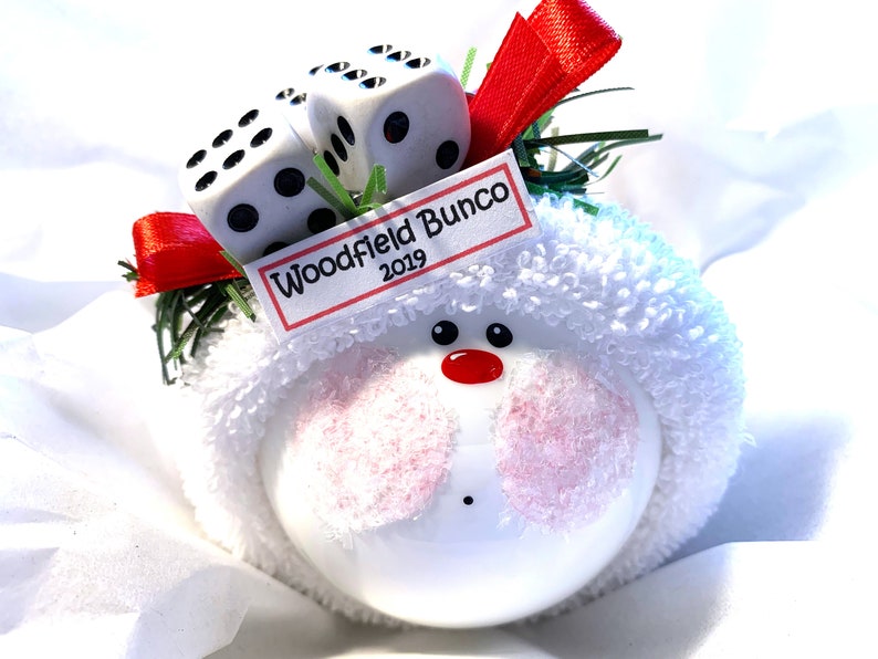 May include: A white ornament with a snowman face, pink cheeks, and a red nose. Two dice sit atop the ornament, along with a tag that reads "Woodfield Bunco 2019". The ornament is decorated with red ribbon and green sprigs.