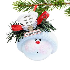 May include: A snowman-shaped Christmas ornament with a white knit hat and rosy cheeks. The ornament has a red nose and black eyes. A heart-shaped tag reads "You left paw prints on our heart." Includes a tag that says "Murphy (year)."