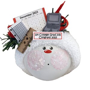 May include: A white snowman ornament with a fluffy hat, pink cheeks, and a red nose. It features a miniature laptop, calendar, notepad, and ruler. The text "1st College Grad job Congrats 2023" is displayed on a sign.