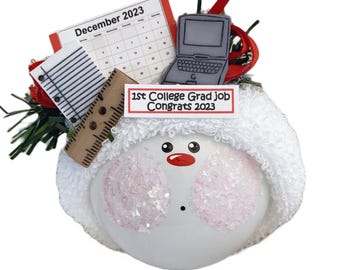 May include: A white snowman ornament with a fluffy hat, pink cheeks, and a red nose. It features a miniature laptop, calendar, notepad, and ruler. The text "1st College Grad job Congrats 2023" is displayed on a sign.