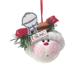 May include: White snowman ornament with a pink face, red nose, and a white knit hat. The ornament has a green sprig of pine, a red ribbon, a black and white grill, and a brown hamburger on top. The ornament has a white tag with the text "Tim (year)" on it.