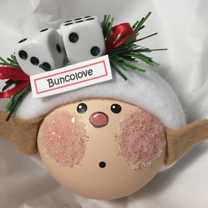 Puede incluir: Un adorno caprichoso con una cara pintada, mejillas rosadas y un sombrero blanco. Dos dados se colocan encima del sombrero, con una cinta roja y el texto "Buncolove" en una peque&ntilde;a etiqueta. El adorno tiene orejas marrones.