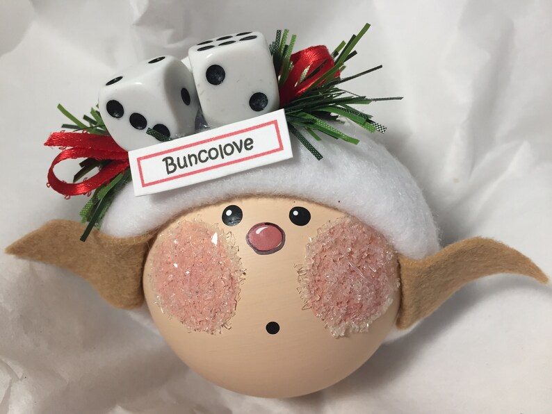 Puede incluir: Un adorno caprichoso con una cara pintada, mejillas rosadas y un sombrero blanco. Dos dados se colocan encima del sombrero, con una cinta roja y el texto "Buncolove" en una peque&ntilde;a etiqueta. El adorno tiene orejas marrones.