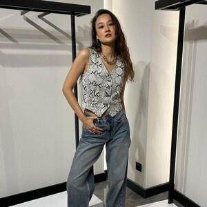Könnte beinhalten: Eine ärmellose Weste mit Schlangenmuster in Schwarz und Weiß, kombiniert mit blauen Jeans. Die Weste hat einen V-Ausschnitt und einen Knopfverschluss. Eine silberne Halskette rundet den Look ab. Das Outfit ist lässig und stilvoll.