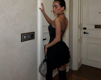 May include: A black dress with a tiered skirt and a halter neckline. The dress has a corset-style bodice and is paired with over-the-knee black boots. A small, silver, beaded handbag is held in the hand.