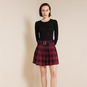 Op de afbeelding: Een zwarte top met lange mouwen gecombineerd met een rood-zwarte geruite geplooide rok. De rok heeft een riem met een rechthoekige gesp. De outfit is een klassieke stijl, geschikt voor diverse gelegenheden.