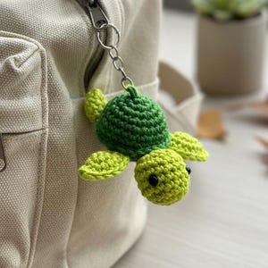 Könnte beinhalten: Ein grüner und gelber gehäkelter Schildkröten-Schlüsselanhänger, der an einer beigefarbenen Canvas-Tasche befestigt ist. Die Schildkröte hat einen strukturierten Panzer und kleine schwarze Augen. Die Tasche hat einen Reißverschluss und eine kleine Tasche.