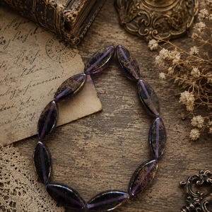 Può includere: Una collana composta da perline ovali viola scuro con inclusioni dorate. Le perline sono infilate insieme, formando un anello. La collana è esposta su una superficie di legno rustico, con elementi vintage sullo sfondo.