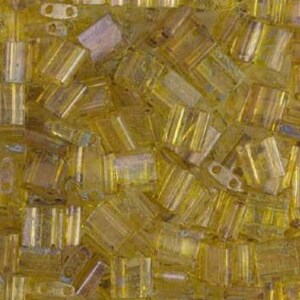 May include: Close-up of a pile of small, rectangular, translucent yellow beads. The beads are scattered and overlapping, creating a textured surface. The beads have a slight shimmer and vary in shade from light to dark yellow.