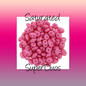 May include: A close-up of a pile of pink SuperDuos beads. The beads are small and have a matte finish. The text "Saturated SuperDuos" is visible at the top and bottom of the image.