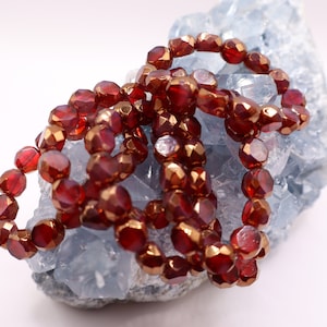 Op de afbeelding: Een streng rode glazen kralen met een gouden metallic afwerking. De kralen zijn gefacetteerd en hebben een licht onregelmatige vorm. De kralen zijn in een losse stapel op een blauw en wit kristal gelegd.