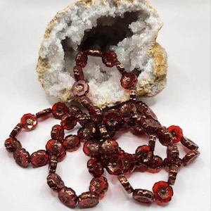 Op de afbeelding: Een streng rode glazen bloemen kralen met een metallic glans. De kralen zijn geregen op een doorzichtige draad en zijn gerangschikt in een los, vloeiend patroon.