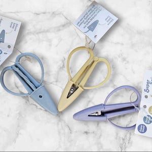 Puede incluir: Tres tijeras pequeñas de colores pastel, azul, amarillo y lavanda, cada una con una funda de seguridad de silicona. Las etiquetas de las tijeras dicen: "Every Handbag needs one. Cuts almost everything. The highest quality, sharpest little shear you will ever own!"