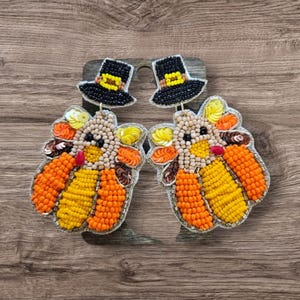 May include: A pair of beaded earrings shaped like turkeys with black pilgrim hats. The turkeys are beaded in shades of orange, yellow, and brown with gold accents.