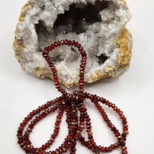 May include: A strand of red garnet beads with a metallic sheen, displayed on a white and brown geode.