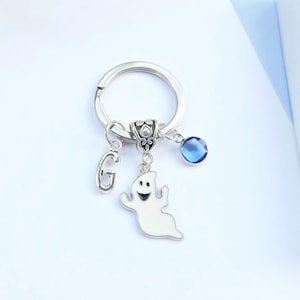 May include: Silver keyring featuring a white ghost charm, a blue crystal bead, and the letter 'G'. The ghost has a smiling face. The background is white and light blue.