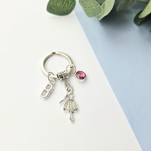 May include: Silver-coloured keyring featuring a ballerina charm, a letter "B" charm, and a pink gemstone. The keyring rests on a white and light blue surface, with green foliage in the background. A delightful accessory.
