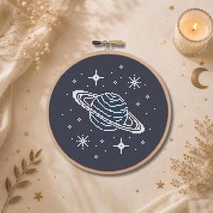 Pode incluir: Um bastidor de bordado circular de madeira contendo um padrão de ponto cruz de um planeta com anéis, feito com fio branco sobre um tecido azul-marinho escuro, rodeado por várias formas de estrelas bordadas em branco no céu noturno.