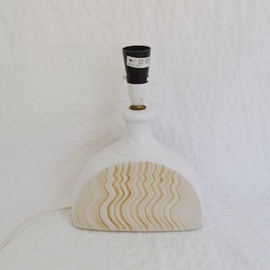May include: A white ceramic table lamp with a black lamp holder. The lamp has a curved base with a beige, wavy pattern. The lamp is on a white surface.
